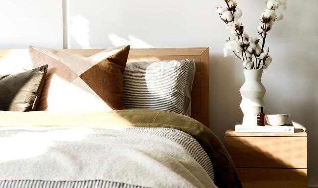 swath of sunlight shines across a well made bed and bedside table with a vase of cotton plants