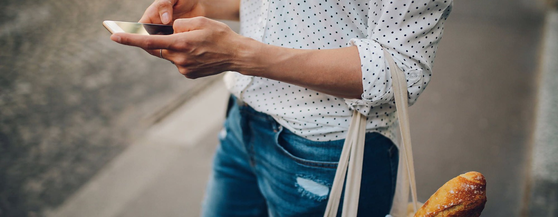 woman walks down the street and texts with a bag of groceries on her arm